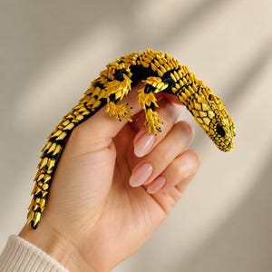 Peut inclure: Une petite figurine de lézard articulée avec un corps noir recouvert de segments pointus jaune vif ressemblant à des écailles. La créature possède une longue queue courbée, quatre pattes avec des griffes visibles et une tête détaillée aux yeux sombres.