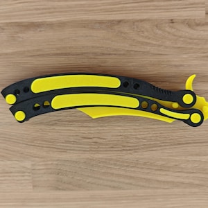 Op de afbeelding: Een gele en zwarte vlindermes, ook wel balisong genoemd, ligt open op een houten oppervlak. Het mes heeft een gele handgreep en lemmet met zwarte details. Het open mes is ongeveer 23 cm lang.
