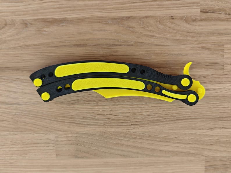 Op de afbeelding: Een gele en zwarte vlindermes, ook wel balisong genoemd, ligt open op een houten oppervlak. Het mes heeft een gele handgreep en lemmet met zwarte details. Het open mes is ongeveer 23 cm lang.