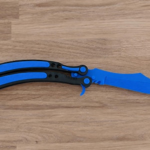 Op de afbeelding: Een blauw en zwart vlindermes, ook wel balisong genoemd, ligt open op een houten oppervlak. Het lemmet en de handvataccenten zijn helderblauw, wat contrasteert met het zwarte handvat. Het mes heeft een gebogen lemmet en een vouwmechanisme.