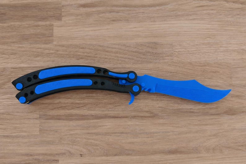 Op de afbeelding: Een blauw en zwart vlindermes, ook wel balisong genoemd, ligt open op een houten oppervlak. Het lemmet en de handvataccenten zijn helderblauw, wat contrasteert met het zwarte handvat. Het mes heeft een gebogen lemmet en een vouwmechanisme.