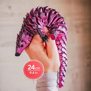 Può includere: Una statuetta di pangolino articolata color magenta con una texture a strati simile a scaglie e dettagli neri sul naso e sugli artigli. Un grafico circolare indica una lunghezza di 24 cm per questo modello animale flessibile e segmentato.