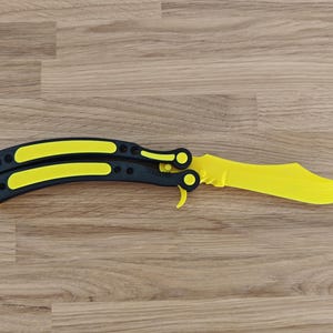 Op de afbeelding: Een gele en zwarte vlindermes, met een gebogen handvat en een gekarteld lemmet. Het handvat is zwart met gele accenten en het lemmet is felgeel. Het mes ligt op een houten oppervlak.