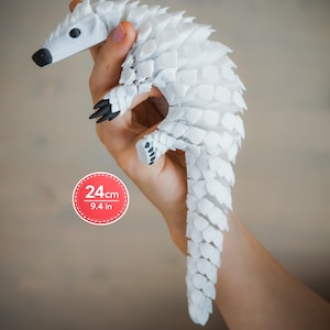 Pode incluir: Uma figura de pangolim articulada branca com garras e olhos pretos, apresentando um design de escamas em camadas. Um gráfico circular vermelho na lateral exibe o texto 24cm e 9.4 in, indicando o comprimento total do objeto.