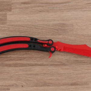 Op de afbeelding: Een rood en zwart vlindermes, ook wel balisong genoemd, ligt open op een houten oppervlak. Het mes heeft een felrood lemmet en zwarte handvatten met rode accenten. Het mes heeft een modern ontwerp.