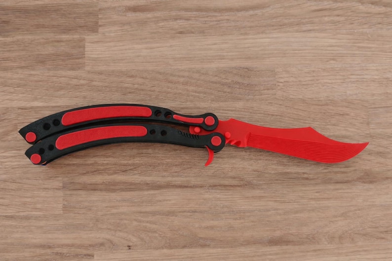 Op de afbeelding: Een rood en zwart vlindermes, ook wel balisong genoemd, ligt open op een houten oppervlak. Het mes heeft een felrood lemmet en zwarte handvatten met rode accenten. Het mes heeft een modern ontwerp.