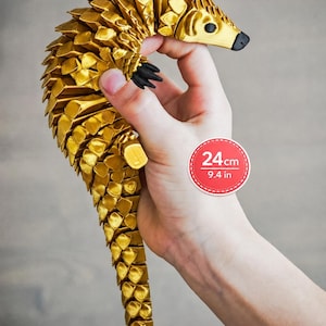 May include: A gold-coloured articulated pangolin figurine featuring a segmented, scale-like body design with black eyes and claws. The figure measures 24 cm in length as indicated by the circular graphic overlay on the image.