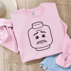 May include: A light pink sweatshirt featuring a black and white graphic of a Lego-style face with a worried expression. The sweatshirt is folded with a white hat and denim shorts in the background.