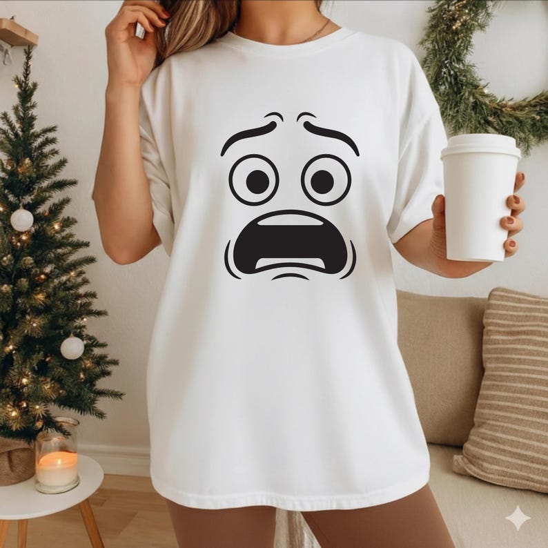 May include: White t-shirt featuring a black cartoon face with wide eyes and an open mouth. The graphic is centered on the shirt. The person is holding a white coffee cup. A small Christmas tree and a wreath are in the background.