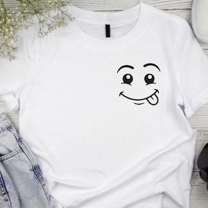 May include: White t-shirt featuring a black cartoon face with eyes, a smile, and a tongue sticking out. The shirt is made of a soft material and has a classic crew neck design. The shirt is a casual wear item.
