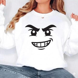 May include: White sweatshirt featuring a black graphic of a cartoon face with an angry expression. The face has large eyes, arched eyebrows, and a wide, toothy grin. The sweatshirt has long sleeves and a crew neck.