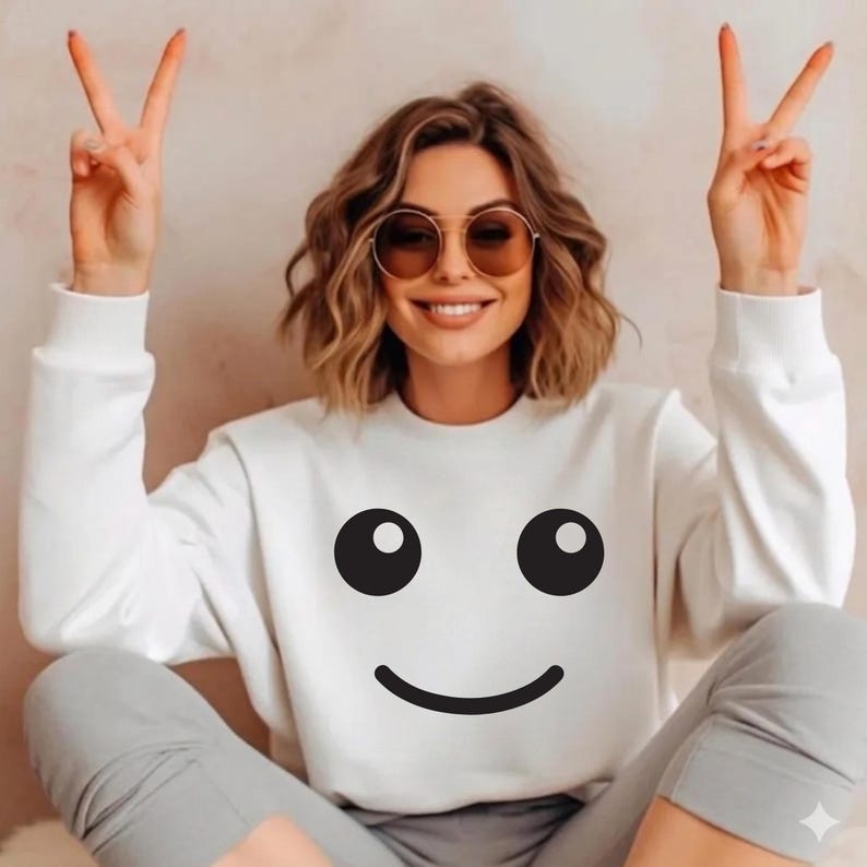 May include: White sweatshirt with a large black smiley face graphic. The sweatshirt is paired with gray pants. The person is wearing round sunglasses and making a peace sign with their fingers.