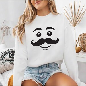 May include: White sweatshirt featuring a black cartoon mustache face design. The graphic includes two large eyes, eyebrows, a mustache, and a smiling mouth. The long-sleeved top is made of a soft material.