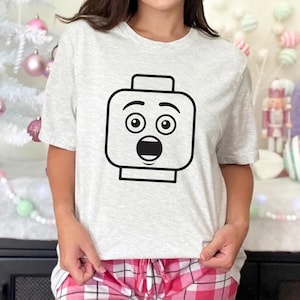 May include: A light gray t-shirt featuring a black graphic of a surprised Lego head. The graphic has large, round eyes and an open mouth. The shirt is short-sleeved and has a crew neck.