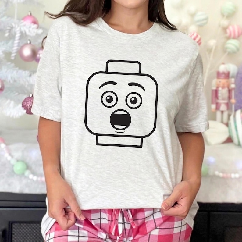 May include: A light gray t-shirt featuring a black graphic of a surprised Lego head. The graphic has large, round eyes and an open mouth. The shirt is short-sleeved and has a crew neck.