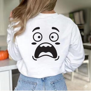 May include: White long-sleeved sweatshirt featuring a large, surprised face graphic on the back. The graphic is a black and white cartoon face with wide eyes and an open mouth. The sweatshirt is worn by a person wearing blue jeans.