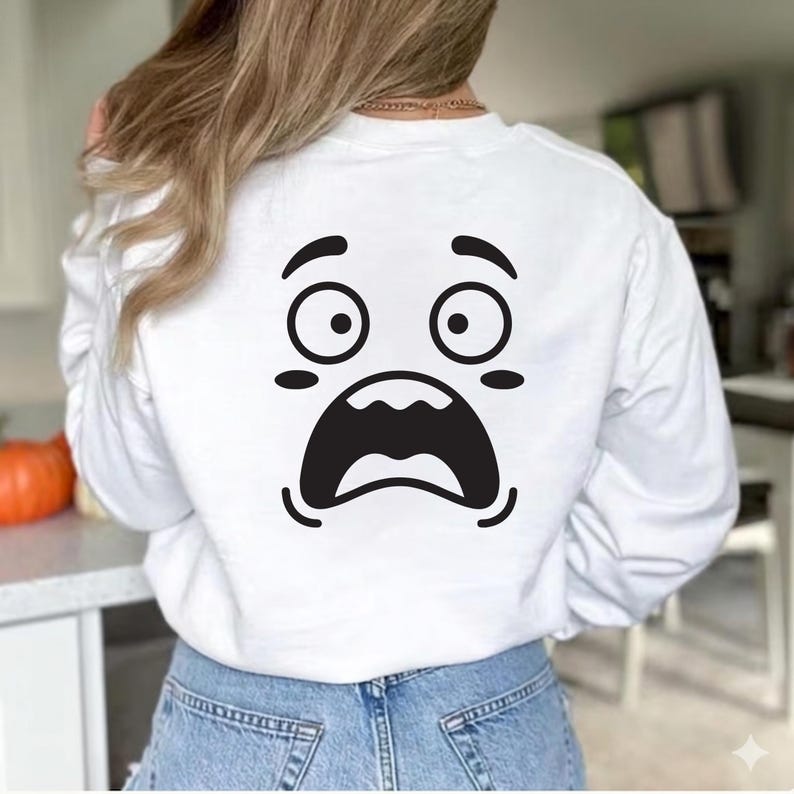 May include: White long-sleeved sweatshirt featuring a large, surprised face graphic on the back. The graphic is a black and white cartoon face with wide eyes and an open mouth. The sweatshirt is worn by a person wearing blue jeans.