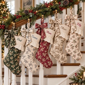 May include: Five personalized Christmas stockings hanging from a wooden staircase railing. Each stocking features a fabric bow, a white eyelet lace cuff, and a name embroidered in script: Dad, Emma, Mom, Oliver, and Lily. The stockings display various patterns including holly, floral prints, and a deer motif.