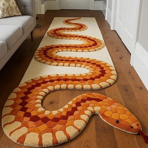 Può includere: Un lungo tappeto decorativo a forma di serpente, con sfondo color crema. Il design del serpente presenta forme geometriche arancioni, rosse e beige. La testa del serpente ha un aspetto strutturato e dettagliato, con una lingua rossa che si estende.
