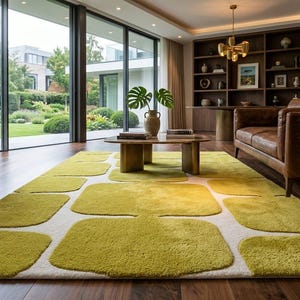 May include: A rectangular area rug featuring a repeating pattern of rounded, olive green squares set against a cream-coloured background. The rug has a textured, plush pile surface and is positioned on a dark wood floor in a living space.