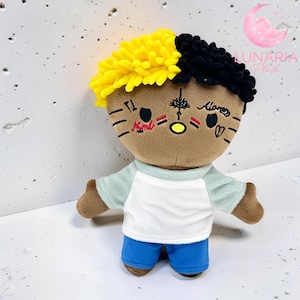 May include: A plush doll with a brown face, yellow and black hair, and facial tattoos. The doll wears a white shirt with light blue sleeves and blue shorts. The doll has open arms and is a cartoon-like character.
