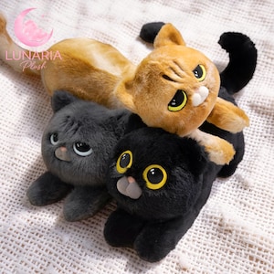 Pode incluir: Três brinquedos de pelúcia em forma de gato, nas cores laranja, cinza e preto, estão dispostos sobre uma superfície branca. O gato laranja está em cima, com o gato cinza embaixo. O gato preto está em primeiro plano. O logotipo Lunaria Plush está no canto superior esquerdo.