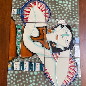 May include: A set of six ceramic tiles arranged to form an abstract image of a person. The artwork features a color palette of white, brown, orange, blue, and red. The tiles are arranged on a wooden surface. The artwork has a unique, artistic style.