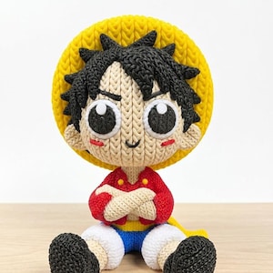 May include: A handmade crochet doll of a cartoon character, featuring a yellow straw hat, black spiky hair, and a red shirt. The doll has large, expressive eyes and is seated with arms crossed. The doll's shorts are blue and white, and the shoes are black.