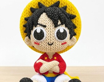 May include: A handmade crochet doll of a cartoon character, featuring a yellow straw hat, black spiky hair, and a red shirt. The doll has large, expressive eyes and is seated with arms crossed. The doll's shorts are blue and white, and the shoes are black.
