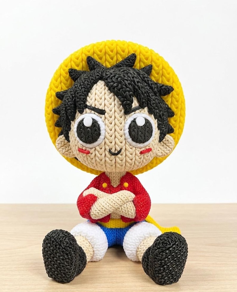 May include: A handmade crochet doll of a cartoon character, featuring a yellow straw hat, black spiky hair, and a red shirt. The doll has large, expressive eyes and is seated with arms crossed. The doll's shorts are blue and white, and the shoes are black.
