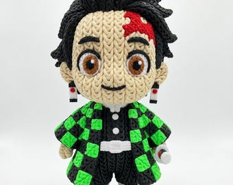 Può includere: Bambola all'uncinetto fatta a mano di un personaggio con capelli neri, occhi marroni e un segno rosso sulla fronte. La bambola indossa una giacca a scacchi nera e verde, pantaloncini neri e scarpe con dettagli rossi. Ha orecchini neri e una benda bianca.