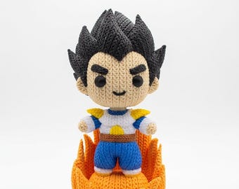 May include: A handmade crochet figure of a character with spiky black hair, wearing a white and blue outfit, standing on an orange flame base. The figure has a neutral expression and is made with yarn.