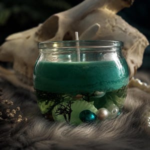 Pode incluir: Uma vela em pote de vidro com uma parte superior de cera verde vibrante. A vela contém elementos decorativos como pequenas folhas, pérolas e uma conta verde-azulado. Um pavio branco se estende. O fundo inclui um crânio e um colar de pérolas.