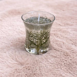 Op de afbeelding: Een heldere glazen kaars met een witte lont, gevuld met een heldere gel en takjes groene kruiden. De kaars staat op een lichtroze, zacht oppervlak. De kaars heeft een uitlopende vorm.