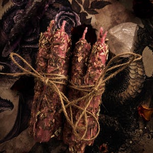 Puede incluir: Un conjunto de velas rosas, adornadas con hierbas secas, atadas con hilo. Las velas se colocan sobre un fondo de ilustraciones florales oscuras y un cristal, creando una estética mística. Las velas miden aproximadamente 15 cm de alto.