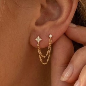 May include: Gold-toned double-piercing earrings. One earring features a square-shaped design with small, clear stones. The second earring is a small, clear stone. Both earrings are connected by delicate gold chains. The earrings are worn on the ear.