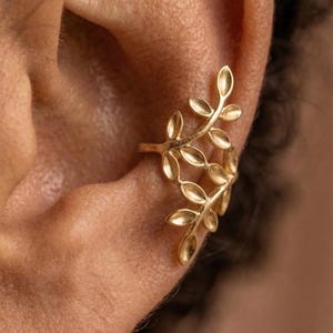 May include: Gold-colored ear cuff featuring a delicate leaf design. The ear cuff wraps around the ear, with multiple leaves arranged to create a natural, organic look. This piece of jewelry is designed to be worn on the ear.