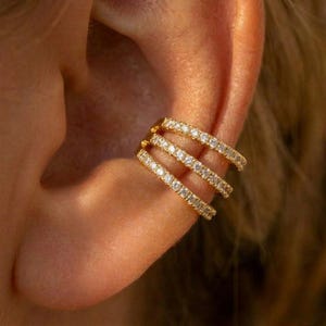 May include: A gold ear cuff featuring three rows of clear, sparkling gemstones. The ear cuff is positioned on the upper ear, with the gemstones arranged in a continuous line along each row. The jewellery has a bright, polished finish.