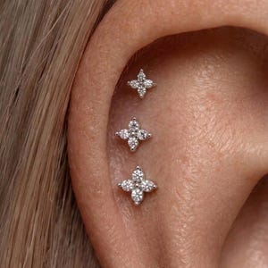 May include: Close-up of an ear with three small, silver-toned stud earrings. Each earring features a four-petal flower design, encrusted with small, sparkling clear stones. The earrings are set in the ear cartilage.