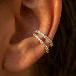 May include: Two gold-colored ear cuffs, each adorned with a row of small, sparkling gemstones. The ear cuffs are positioned on the ear, creating a layered effect. The image is a close-up, highlighting the jewelry's details.