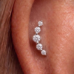 Op de afbeelding: Een gebogen oormanchet met vijf ronde, heldere edelstenen in een zilverkleurig metaal. De edelstenen nemen in grootte af. De sieraden worden op het oor gedragen.