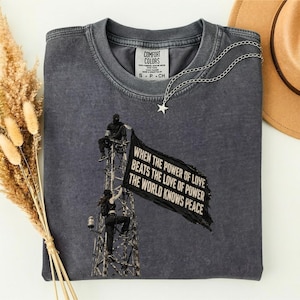 Può includere: Una t-shirt a maniche corte color grigio antracite con una grafica raffigurante due figure che si arrampicano su una torre di metallo e tengono una bandiera nera. La bandiera riporta il testo: WHEN THE POWER OF LOVE BEATS THE LOVE OF POWER THE WORLD KNOWS PEACE.