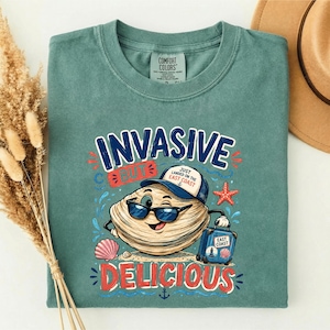 May include: A light green t-shirt featuring a graphic of a cartoon clam wearing sunglasses and a blue cap, holding a blue suitcase. The text reads "INVASIVE BUT DELICIOUS" and "JUST LANDED ON THE EAST COAST" with a starfish and seashell accents.