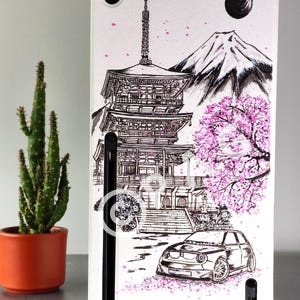 以下が含まれることがあります： 日本の五重塔、富士山、車が黒いインクで描かれた白い長方形のゲーム機。デザインにはピンクの桜のアクセントと暗い円形の月が含まれています。コンソールには黒い垂直のアクセントが施されています。