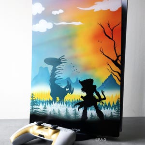 Könnte beinhalten: Eine individuell bemalte PlayStation 5-Konsole mit passendem Controller. Die Konsole zeigt eine lebendige Landschaft im Sonnenuntergang mit Silhouetten. Der Controller ist weiß mit goldenen Akzenten. Das Kunstwerk zeigt einen Krieger und ein Wesen.