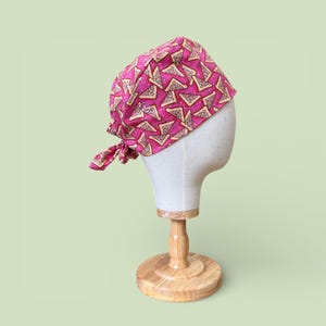 May include: A pink surgical scrub cap featuring a repeating pizza slice design. The cap is tied at the back and displayed on a mannequin head. The background is a light green colour.