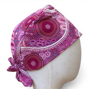 May include: A magenta and white patterned head covering, tied at the back. The fabric features a circular design with dots and floral elements in shades of magenta, pink, blue, and white. The head covering is displayed on a mannequin head.
