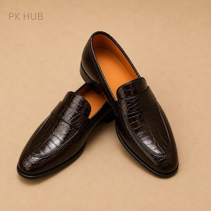 May include: A pair of dark brown loafers with a glossy crocodile-embossed texture and a smooth orange interior lining. The shoes feature a classic slip-on design with a strap across the vamp. The text PK HUB is visible in the upper left corner.