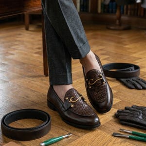 May include: Brown leather loafers with woven detailing and gold-colored hardware. The shoes are paired with a matching belt, gloves, and a pen. The loafers are a classic style, suitable for formal or business attire.