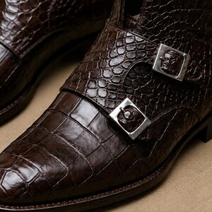 May include: Close-up of a pair of brown leather boots with a crocodile skin texture. The boots feature silver buckles with a skull design. The leather has a glossy finish, and the boots appear to be new.
